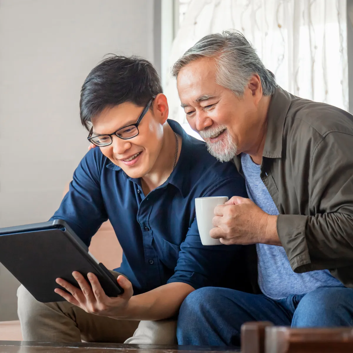Elderly man with coffee cup with arm around younger man helping him with computer